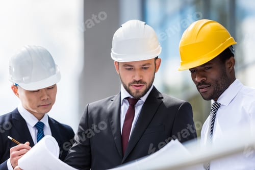Preview: Multiethnic group of professional architects in helmets working with blueprint outside modern