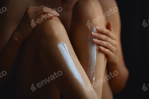 Preview: Woman applying white nourishing cream to both shins of her leg. Close-up of shins and knees