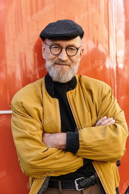 Preview: Dapper senior man stands with arms crossed, exuding charm in an eye catching jacket and trendy cap.