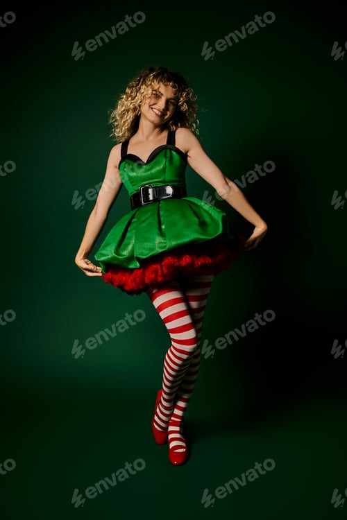 Preview: jolly woman in new year elf costume touching hem of her green dress posing on dark green backdrop