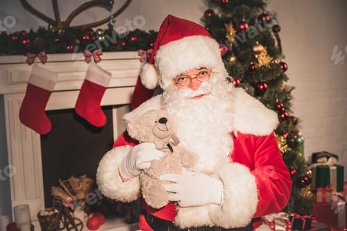 Preview: santa claus holding teddy bear and looking at camera while sitting near fireplace
