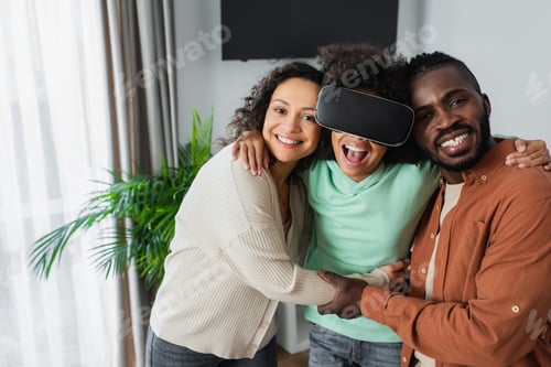 Preview: happy african american parents hugging excited preteen daughter gaming in vr headset