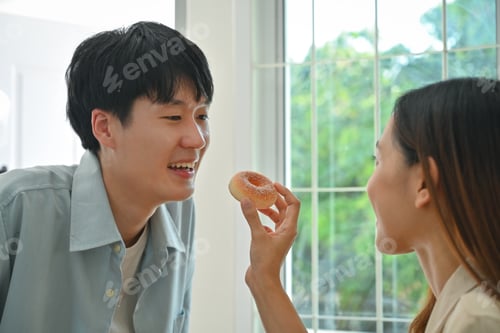 Preview: Young woman feeding his boyfriends with donut while cooking in modern kitchen at home.
