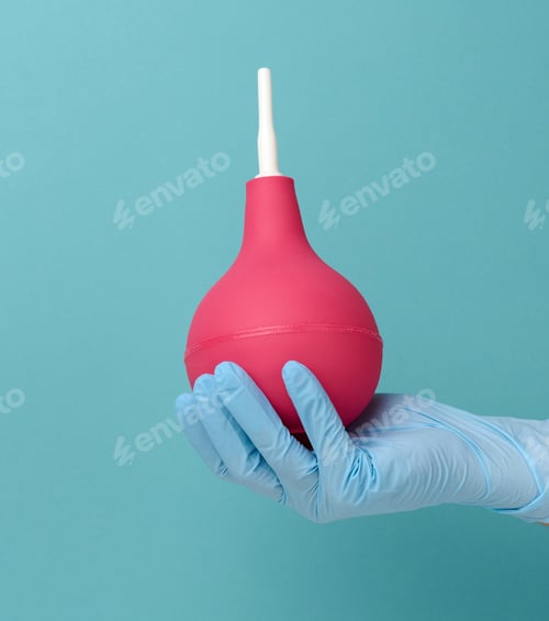 Preview: Hand in a blue medical glove holds a pink rubber enema on a blue background
