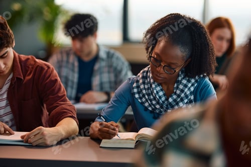 Preview: Black college student writing in her notebook while studying in the classroom.