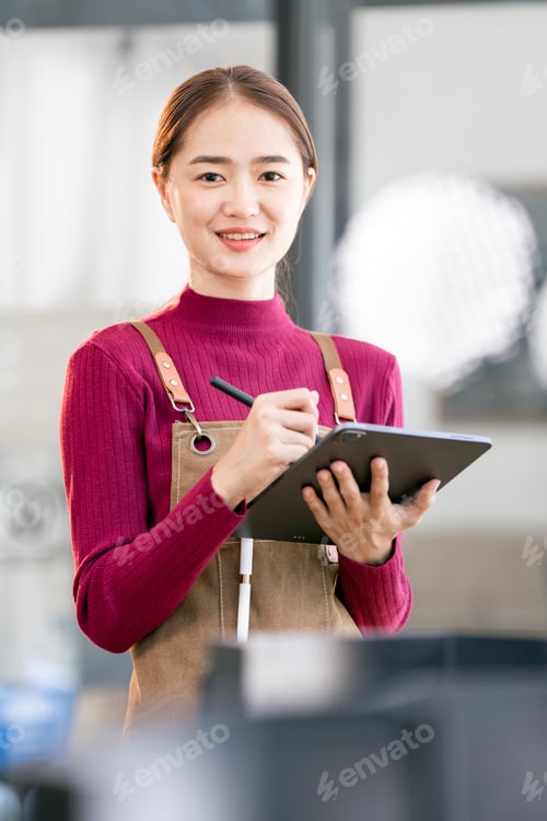 Preview: Successful small business owner in casual wearing brown apron holding tablet standing at entrance