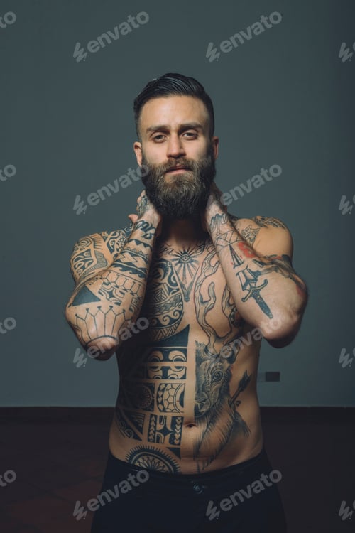 Preview: Portrait of young man with beard, bare chest covered in tattoos, hands behind neck