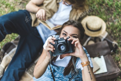 Preview: top view of woman taking photo with vintage camera while relaxing in park with boyfriend