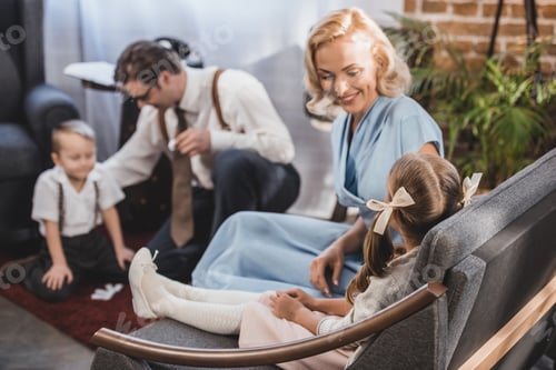 Preview: happy mother looking at cute little daughter sitting on sofa while father with son playing dominoes