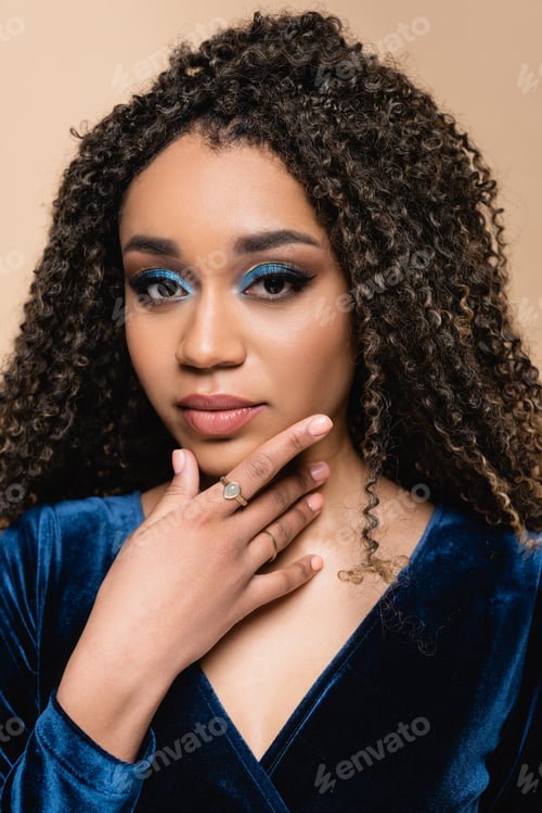 Preview: curly african american woman with makeup touching chin isolated on beige