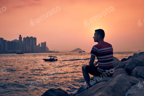 Preview: Young man sitting on stones against Hong Kong cityscape with skyscrapers at golden sunset.