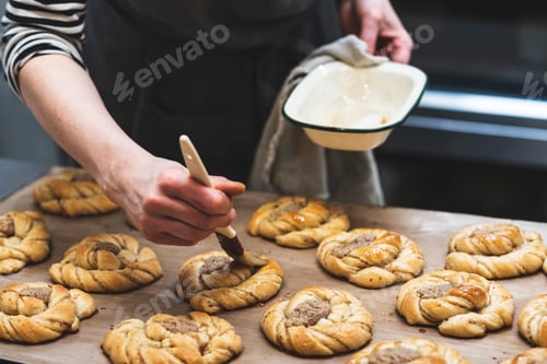 Preview: Baker with a pastry brush adding glaze to a tray of cinnamon buns ready for the oven.