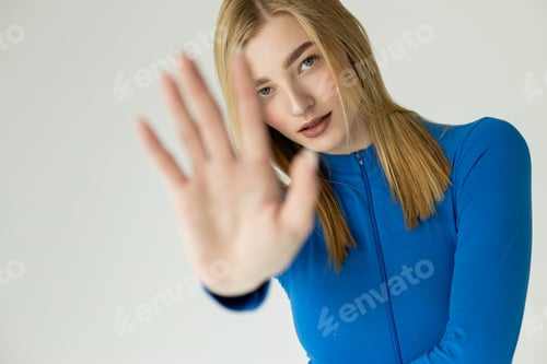 Preview: young woman in white turtleneck showing stop gesture with blurred outstretched hand isolated on grey