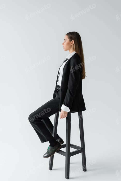 Preview: side view of young brunette woman in black suit with tie sitting on wooden high chair on grey