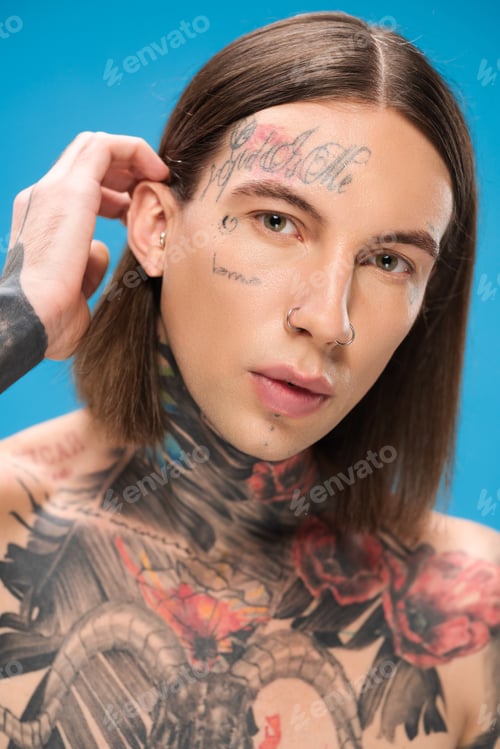 Preview: young and pierced man with tattoos looking at camera while adjusting hair isolated on blue