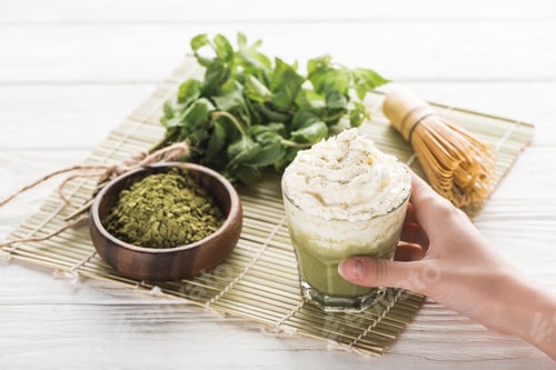 Preview: cropped view of woman holding matcha matcha tea with whipped cream on table with whisk, powder and