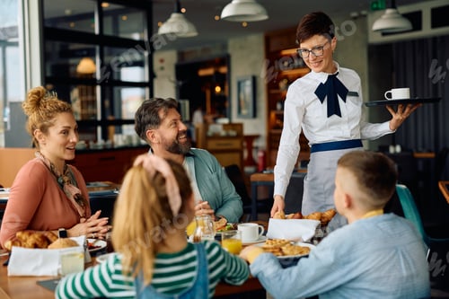Preview: Happy waitress talking to a family while serving them during breakfast at hotel restaurant.