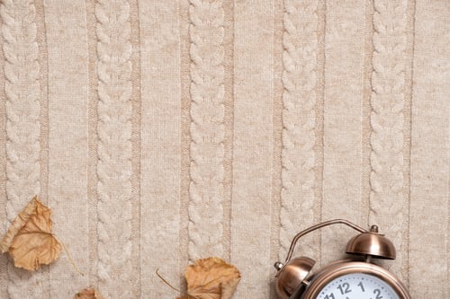 Preview: Alarm clock and dry leaves on a cozy beige sweater. Wellness time concept. Top view. Copy space