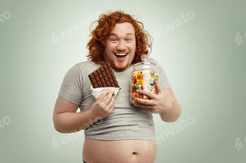Preview: Portrait of happy and joyful red-haired male wearing undersized t-shirt standing at blank studio wal