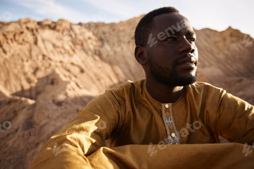 Preview: African American Man Sitting on Sand Dune in Desert and Looking Away in Sunlight