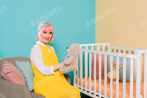 Preview: happy retro styled pregnant pin up woman with pink hair sitting with teddy bear near baby cot
