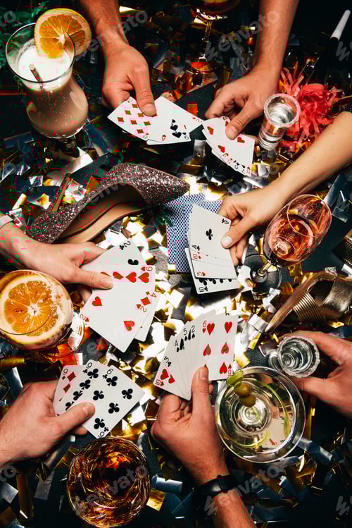 Preview: cropped shot of friends with alcoholic cocktails playing poker at table covered by golden confetti