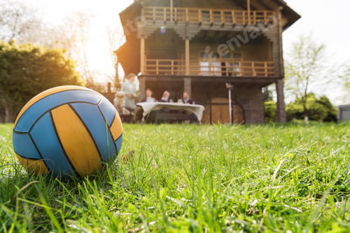 Preview: close-up view of leather ball on green grass and family spending time together near country house