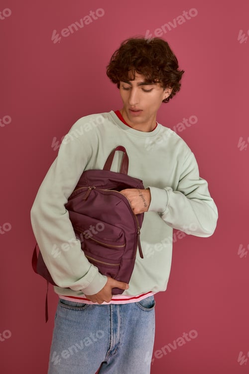 Preview: Stylish young man in pastel sweater interacts with backpack against a vibrant pink background