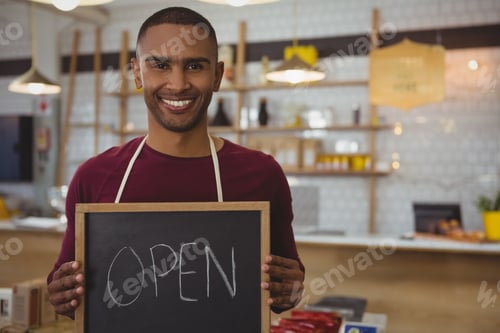 Preview: African American man wearing apron holding OPEN chalkboard sign at wooden counter in coffee shop