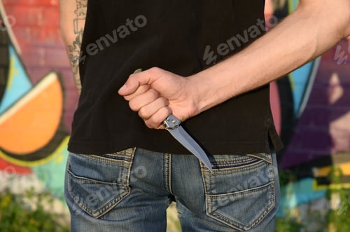 Preview: Back view of young caucasian man with knife in his hand against ghetto brick wall with graffiti