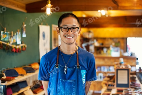 Preview: Japanese man wearing blue apron and glasses standing in a leather shop, smiling at camera.