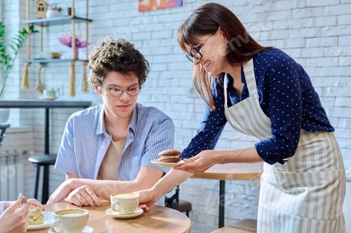 Preview: Woman in an apron with cup of coffee and plate of cake serving young male in cafeteria