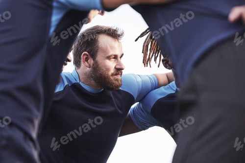 Preview: Below caucasian rugby player standing in a huddle with his teammates outside on a field. Young male