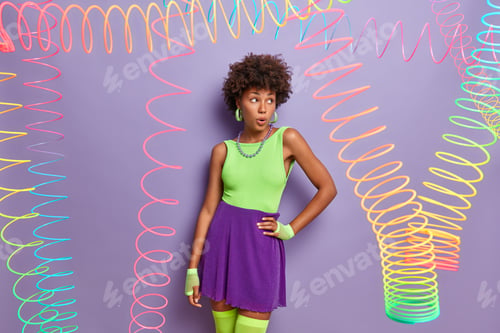 Preview: Surprised dark skinned woman keeps hand on waist, looks shocked away, wears bright shirt, skirt and