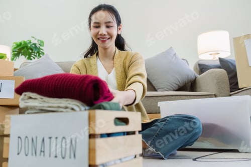 Preview: Asian young woman packing clothes at home, putting on stuff into donate box with second hand clothes