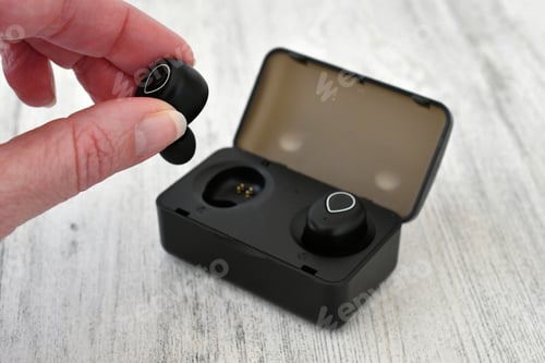 Preview: A woman picking up a black wireless earbud with a charging case in the background