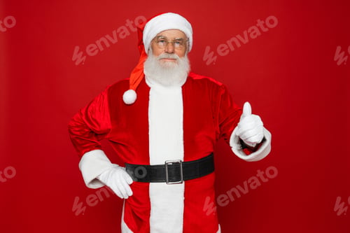 Preview: Santa Claus showing thumb up hand like gesture looking at camera isolated on red background.