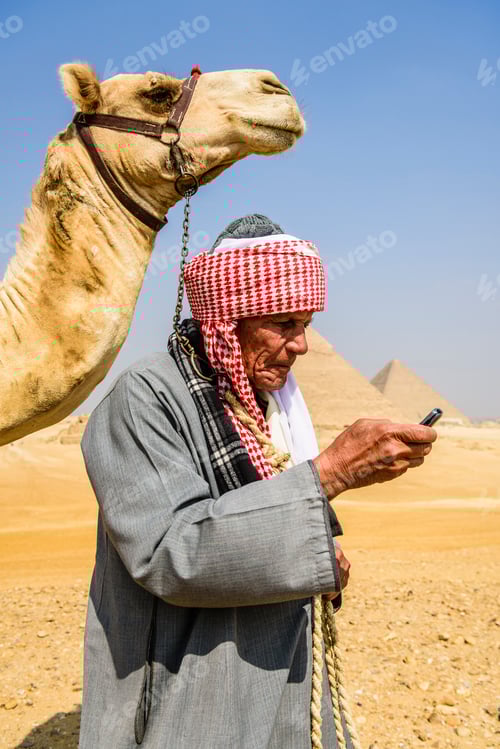 Preview: A guide in traditional Arab dress, robe and turban using a mobile phone, and his camel.