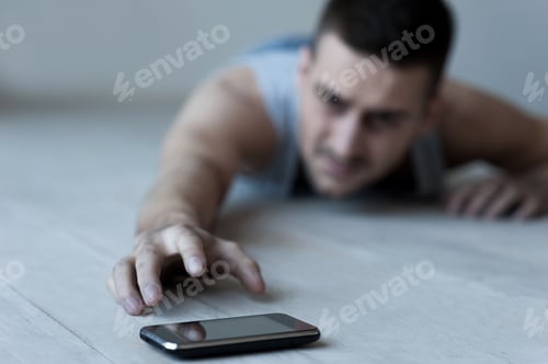 Preview: Help me! Frustrated young man lying on the floor and stretching hand to the mobile phone