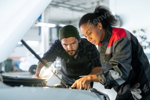 Preview: Young bearded man in workwear holding lamp and bending over open hood