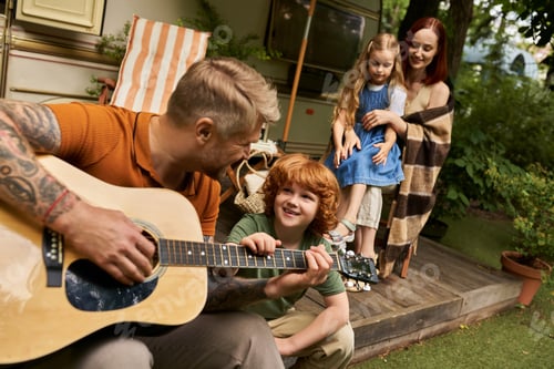 Preview: tattooed man playing acoustic guitar to cheerful redhead kid near family and modern trailer home