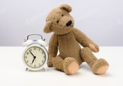 Preview: Brown teddy bear sits on a white table and a round alarm clock