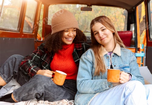 Preview: Two happy multiracial girlfriends on car trunk in fall forest, embracing and drinking hot tea
