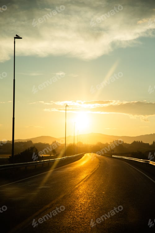 Preview: Golden sunset over a winding road with streetlights and mountains in the background