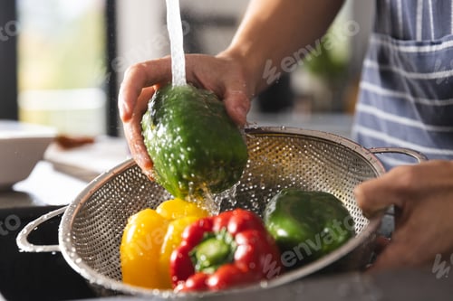 Preview: Midsection of biracial man in apron washing peppers in kitchen sink