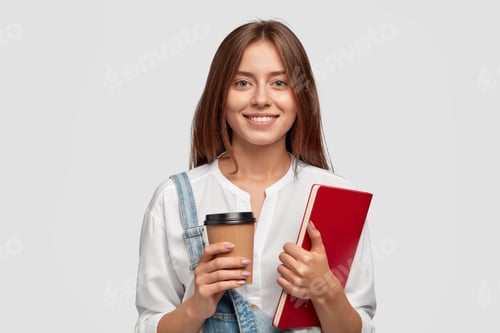 Preview: Cheerful happy woman with toothy smile, carries takeaway coffee and red book, glad to finish studyin