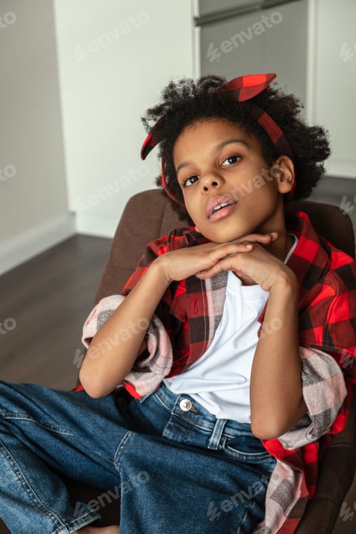 Preview: Cute African-American girl in a red shirt and jeans sitting on a chair at home.Diverse people.