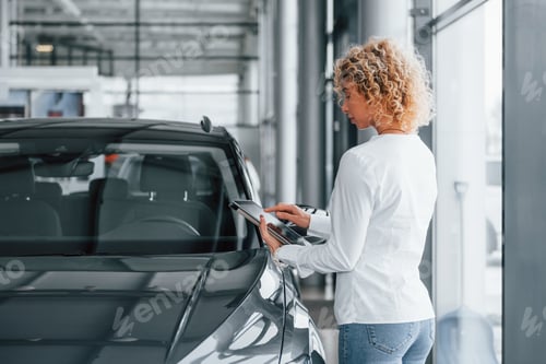 Preview: Holding tablet in hands. Woman with curly blonde hair is in autosalon