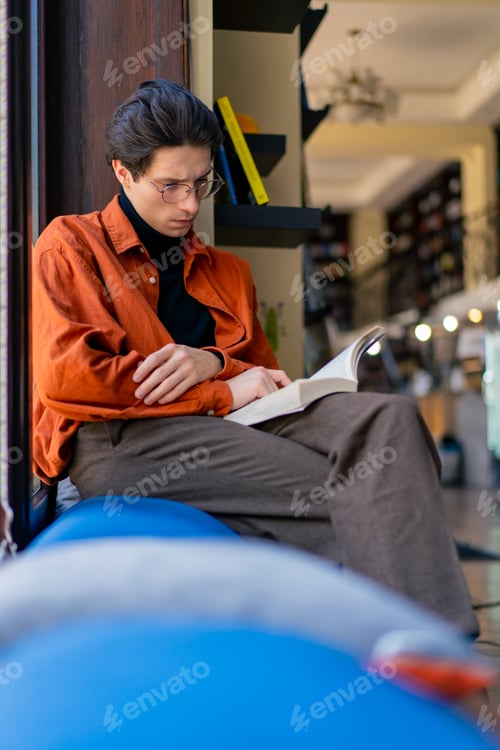 Preview: a young guy with glasses is sitting in library cafe and carefully reading a book about hobbies