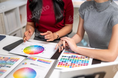 Preview: Close up of woman holding pencil talking to coworker over color chart as sitting at table in office.